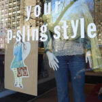 Window display featuring a mannequin dressed in jeans and a lace top with a Psling, reflected cityscape in the glass, and “your p-sling style” text on the window.