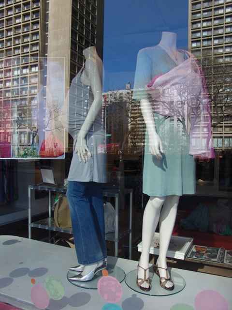 Storefront display with two mannequins, one styled with a Psling baby carrier in a pastel outfit, reflecting the surrounding New York City buildings.