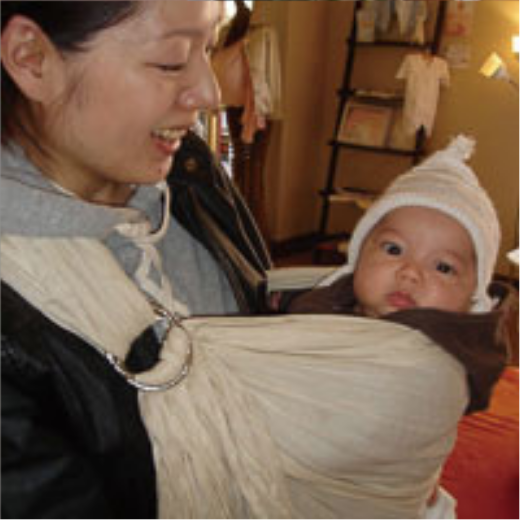 ニット帽をかぶった赤ちゃんをアイボリーのスリングで抱っこする笑顔の女性。
