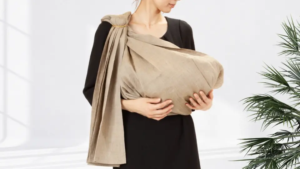 ベージュのピースリングで赤ちゃんを抱く女性の写真。テキスト「赤ちゃんを感じて、抱きしめる。スリングが生まれた理由と、心と身体にやさしい構造。About Sling | What Makes PsLing Special」が重ねられている。